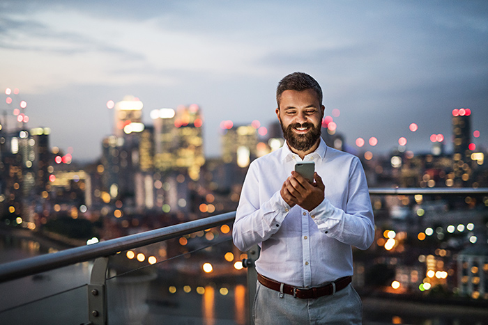 smiling man uses a cell phone on a balcony that overlooks a lighted cityscape
